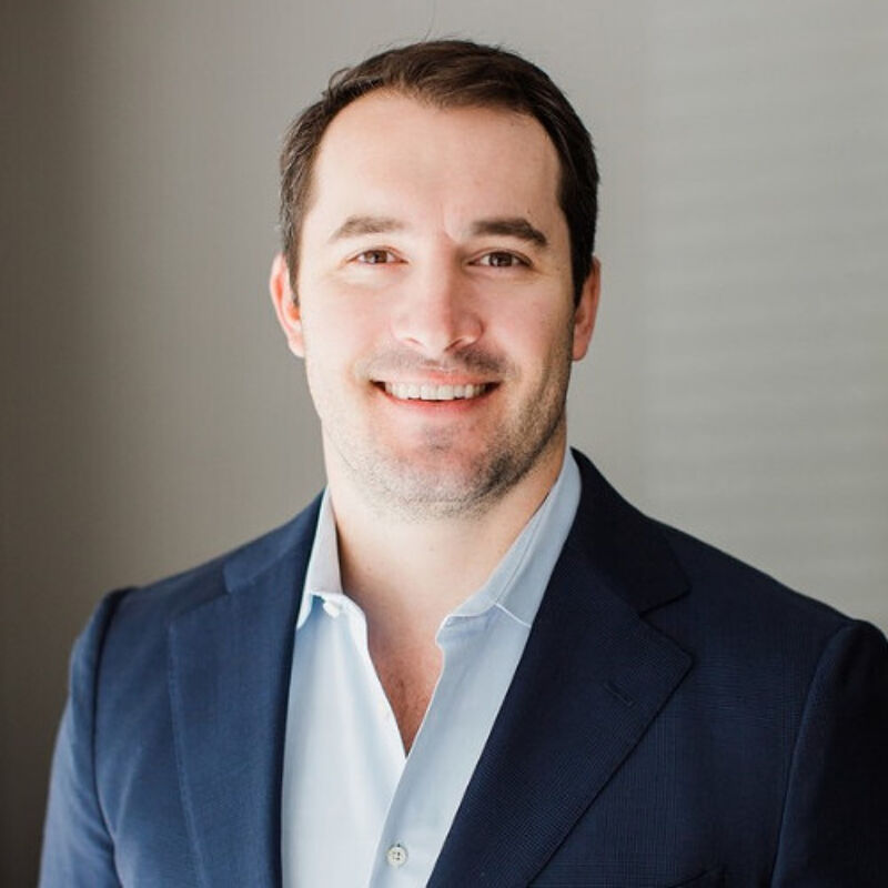The image shows a man with short brown hair and a light complexion. He is wearing a navy blue blazer over a light blue dress shirt. He has a slight smile and appears to be in his late 20s or early 30s. The background is a blurry gray, suggesting an indoor setting.
